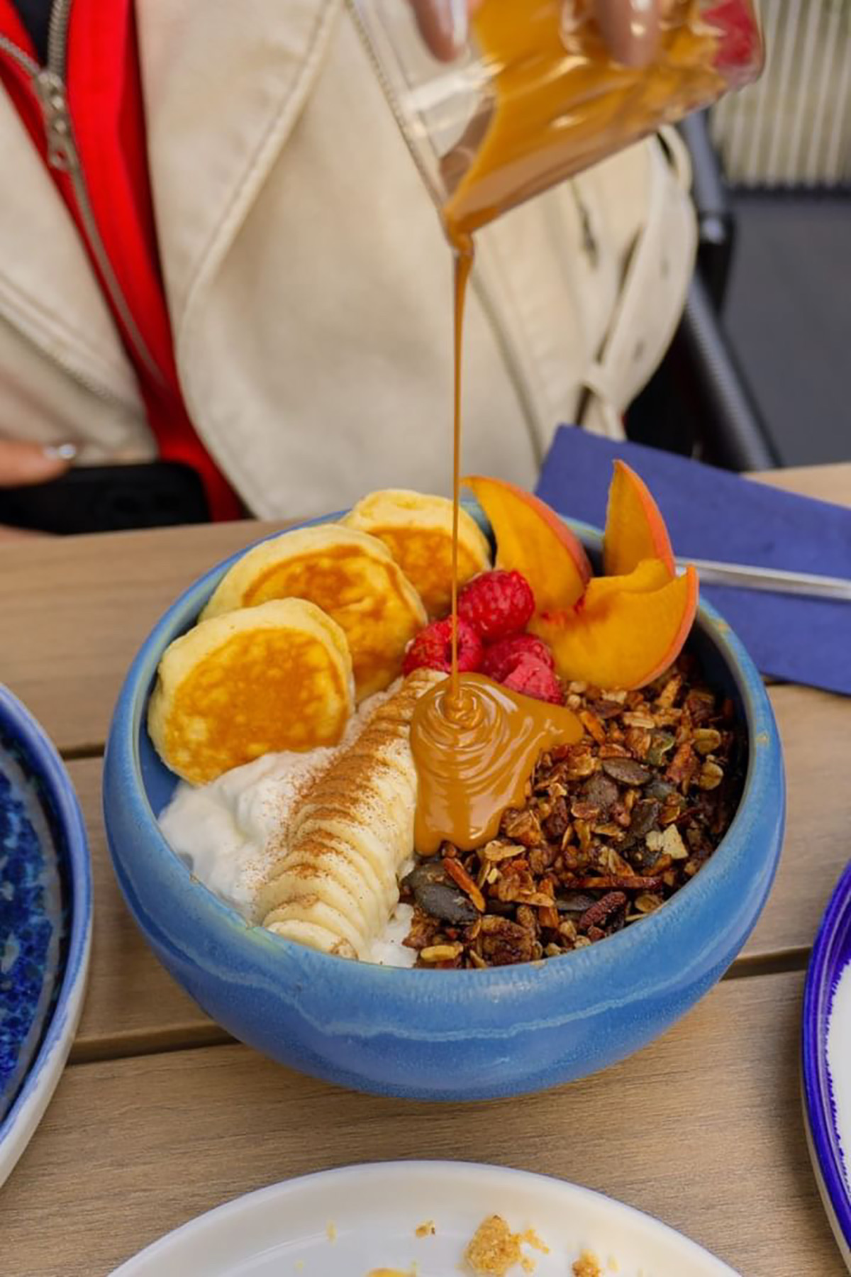 „Cloud Kitchen“: In einer blauen Schale ist Joghurt getoppt mit Granola, Mini Pancakes, Banane, Beeren und Kaki. Von oben wird eine braune Sauce in die Schale geleert.