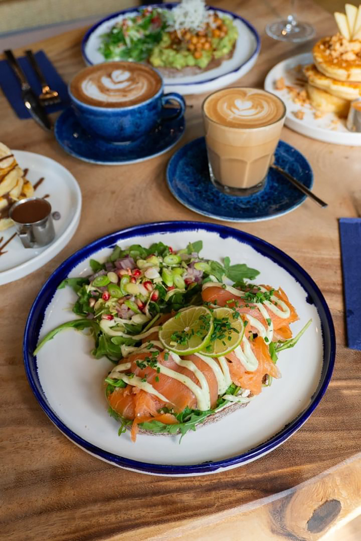 Auf einem weißen Teller mit blauem Rand liegt im „Cloud Kitchen“ ein Brot mit Lachs und einem Beilagensalat, am oberen Bildrand steht ein Kaffee im Glas auf einer blauen Untertasse.
