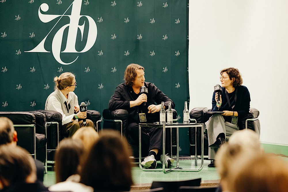 Drei Personen im Panel-Talk auf der ADC Design Conference, sitzend in schwarzen Ledersesseln mit Mikrofonen vor grünem ADC-Bühnenhintergrund.