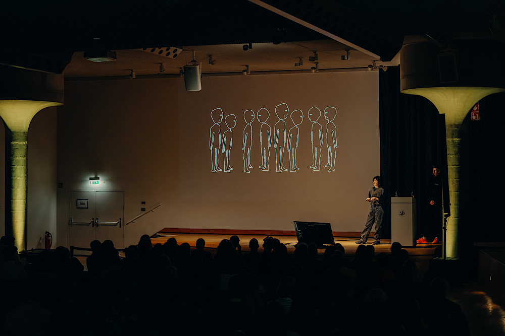 Speakerin auf der Bühne der ADC Design Conference, im Hintergrund acht stilisierte blaue Figuren-Illustrationen auf Leinwand, Publikum im Vordergrund.