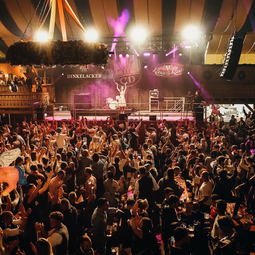 Bühnenblick von oben auf das brechend volle WELLER'S Klauss & Klauss Dinkelacker Festzelt beim Cannstatter Wasen, Sängerin performt vor jubelnder Menge mit erhobenen Armen unter der Zeltdecke.