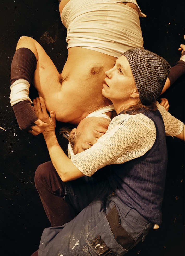 Zwei Schauspieler:innen liegen eng umschlungen am Boden der Bühne, eine Frau mit Wollmütze hält einen Mann mit nacktem Oberkörper im Arm.