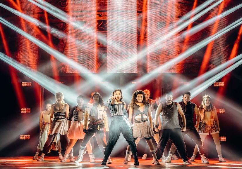 Szene aus dem Pop-Musical „& JULIA“: Julia steht in Jumpsuit und Korsagenoberteil im Zentrum der Bühne, die Faust erhoben, umgeben vom Ensemble in Kostümen aus Renaissance-Mode und Streetwear. Gekreuzte Lichtstrahlen und eine rot leuchtende Bühnenwand rahmen die Gruppe.