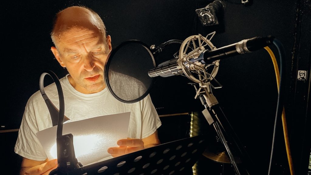 Orlando Schenk liest bei der Aufnahme-Session für Struktur Stammheim ein Skript vor einem Mikrofon, beleuchtet von einer Leselampe im abgedunkelten Studio.