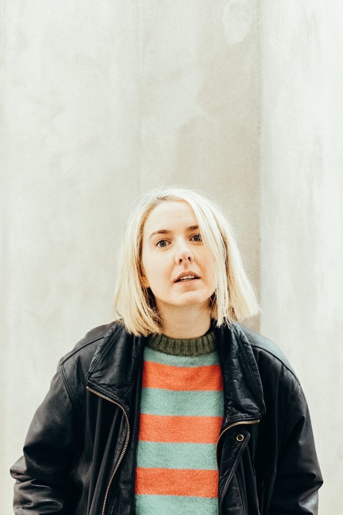 Laura Gertken, Dozentin der Pop-Webinare 2026, mit blondem Bob, schwarzer Lederjacke und gestreiftem Pullover vor einer hellen Betonwand
